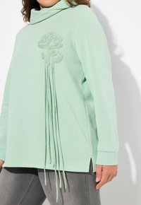 Top lungo a maniche con collo a cowl color verde menta, presenta un design ricamato e filamenti di tessuto allungati sul davanti. Fessure laterali all'orlo.