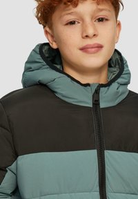 Gepolsterte Jacke mit Kapuze, schwarzem oberen Teil und türkisfarbenem unteren Teil. Reißverschluss mit kontrastierenden Nähten.