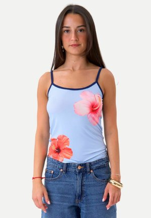 Młoda kobieta z długimi ciemnymi włosami, ubrana w jasnoniebieski tank top z różowymi kwiatowymi nadrukami oraz niebieskie, wysokie spodnie dżinsowe na jednolitym tle.