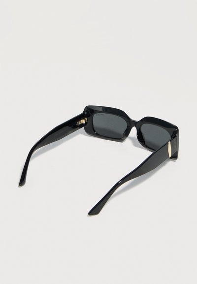 Μαύρα ορθογώνια γυαλιά ηλίου με σκουρόχρωμους φακούς και ελαφρώς κυρτά πλαϊνά, απεικονίζονται πάνω σε απλό ανοιχτό φόντο.