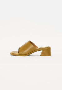 Brown leather heeled mules with an open toe, featuring a V-shaped cutout and a wide strap across the foot. Sturdy block heel design.