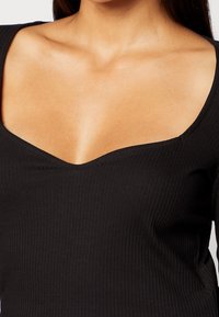 Musta ribbattu toppi, jossa on leveä V-pääntie ja pitkät hihat. Valmistettu sileästä materiaalista, jossa on vartalonmyötäinen muotoilu ja pehmeä tekstuuri.