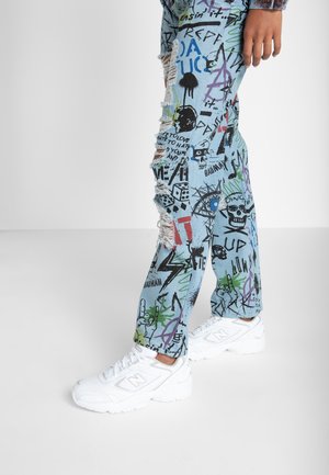Nogi w jasnoniebieskich dżinsach z kolorowymi nadrukami w stylu graffiti i przetarciami, połączone z białymi butami sportowymi New Balance na jednolitym tle.