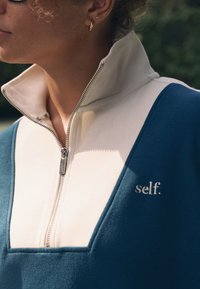 Felpa con zip color teal e crema, caratterizzata da un colletto alto, tessuto morbido e logo "self." ricamato sul petto, con dettaglio della cerniera argentata.