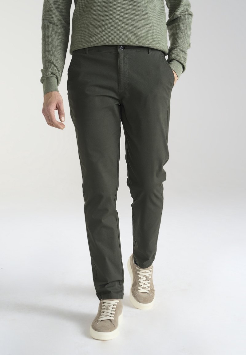 Homme portant un pantalon vert foncé, un pull vert clair et des baskets beiges, marchant avec une main dans la poche.