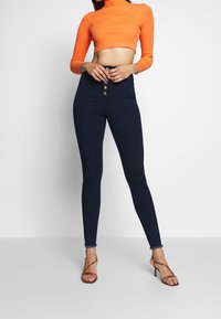 Vaqueros de tiro alto en color azul marino con cierre de cinco botones, combinados con un top de punto corto de color naranja con cuello de tortuga. La modelo lleva sandalias rojas con tiras.