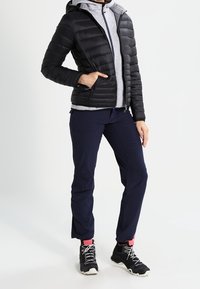 Zwarte pufferjack met capuchon over een grijze zip-up hoodie, marineblauwe broek en zwarte sneakers met witte zolen. Bevat gestructureerde materialen en ritsen.