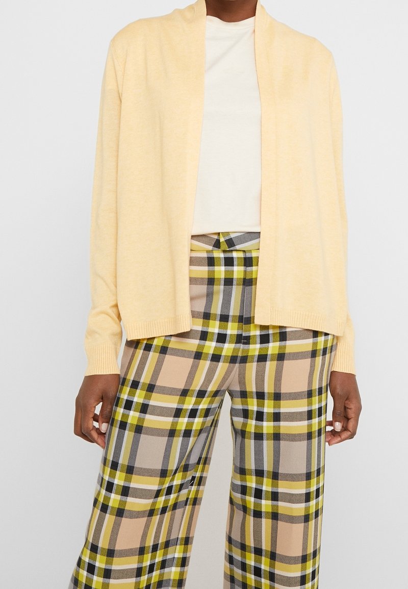 Cardigan giallo chiaro con apertura frontale e polsini a costine, abbinato a pantaloni a gamba larga scozzesi nei colori beige, verde, grigio e bianco.