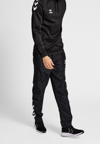 Sudadera atlética negra con cordón, que presenta un logo. Combinada con pantalones de chándal negros, destacados por rayas blancas y puños elásticos. Zapatillas negras.