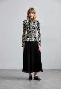 Zwart en crèmekleurig gestreepte top met lange mouwen, gecombineerd met een zwarte geplooide maxi-rok, afgemaakt met zwarte loafers. Gevoelige details van de stof zichtbaar.