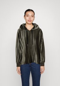 Felpa in velluto verde scuro con pieghe verticali, ampio colletto e lacci. Maniche lunghe con polsini elastici. Abbinata a jeans blu.