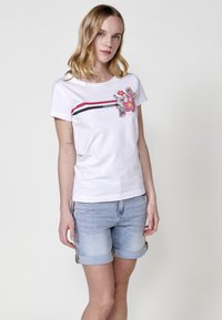 Camiseta blanca de algodón con manga corta, que presenta un diseño floral colorido y franjas horizontales rojas y azul marino en el pecho.