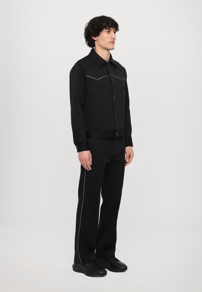 Black jacket with turquoise stitching, button closure, and a collar, paired with matching trousers featuring side stripes and black sneakers.