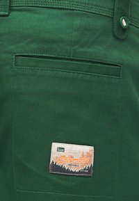 Πράσινη τσέπη παντελονιού από ύφασμα με λουράκι ζώνης που κλείνει με κουμπί και τετράγωνο μπαλώμα με το λογότυπο του Columbia Sportswear Co. ραμμένο κοντά στο κάτω μέρος.