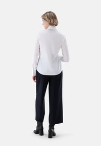 Camicia bianca a maniche lunghe con colletto sul retro, abbinata a pantaloni navy a gamba larga e stivaletti neri con tacco, vista da dietro.