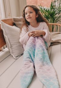 Odeja v obliki sirene v mehkih, pastelnih barvah z vzorcem lusk. Topel, puhast material se tesno prilega otroku, ki sedi na postelji.