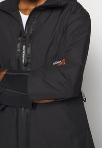 Veste imperméable noire avec fermeture éclair frontale, tissu texturé et marques visibles sur le côté et la poitrine. Comprend une capuche et des poignets réglables.