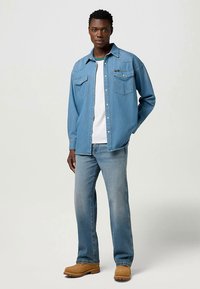 Šviesiai mėlyna džinsinė marškinėliai su dviem krūtinės kišenėmis, dėvimi ant balto marškinėlio, derinami su išblukusiais mėlynais džinsais ir rudu avalyniu.