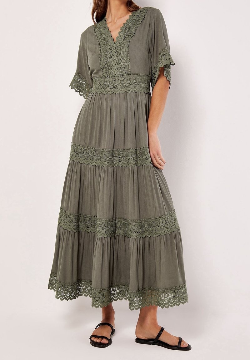 Femme portant une longue robe vert olive avec des détails en dentelle sur les manches, le décolleté, la taille et la jupe à volants, associée à des sandales noires.