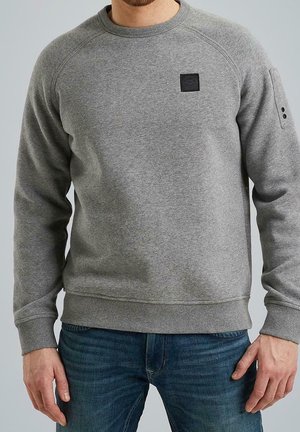 Mann trägt ein schlichtes graues Sweatshirt mit Rundhalsausschnitt und Schulterpatch sowie blaue Jeans, steht vor einem einfarbigen Hintergrund.