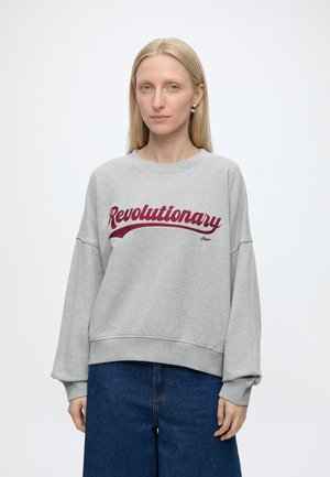 Femme aux longs cheveux blonds portant un sweat gris avec "Revolutionary" en écriture rouge et un jean bleu sur fond blanc.