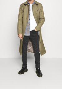 Olijfgroene trenchcoat met dubbelzijdige knopen, grijze geruite shirt daaronder, zwarte spijkerbroek en glanzende zwarte enkellaarsjes.