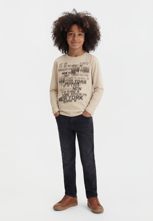 Barn med krøllete hår som har på seg en beige genser med lange ermer med teksten New York, svarte jeans og brune joggesko, står med hendene i lommene.