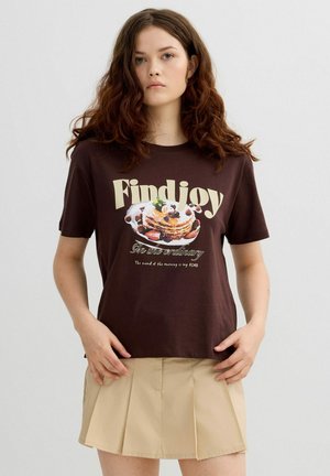 Joven con cabello largo y rizado que lleva una camiseta marrón con un diseño de pancakes con bayas y una falda plisada beige, posando frente a un fondo liso.