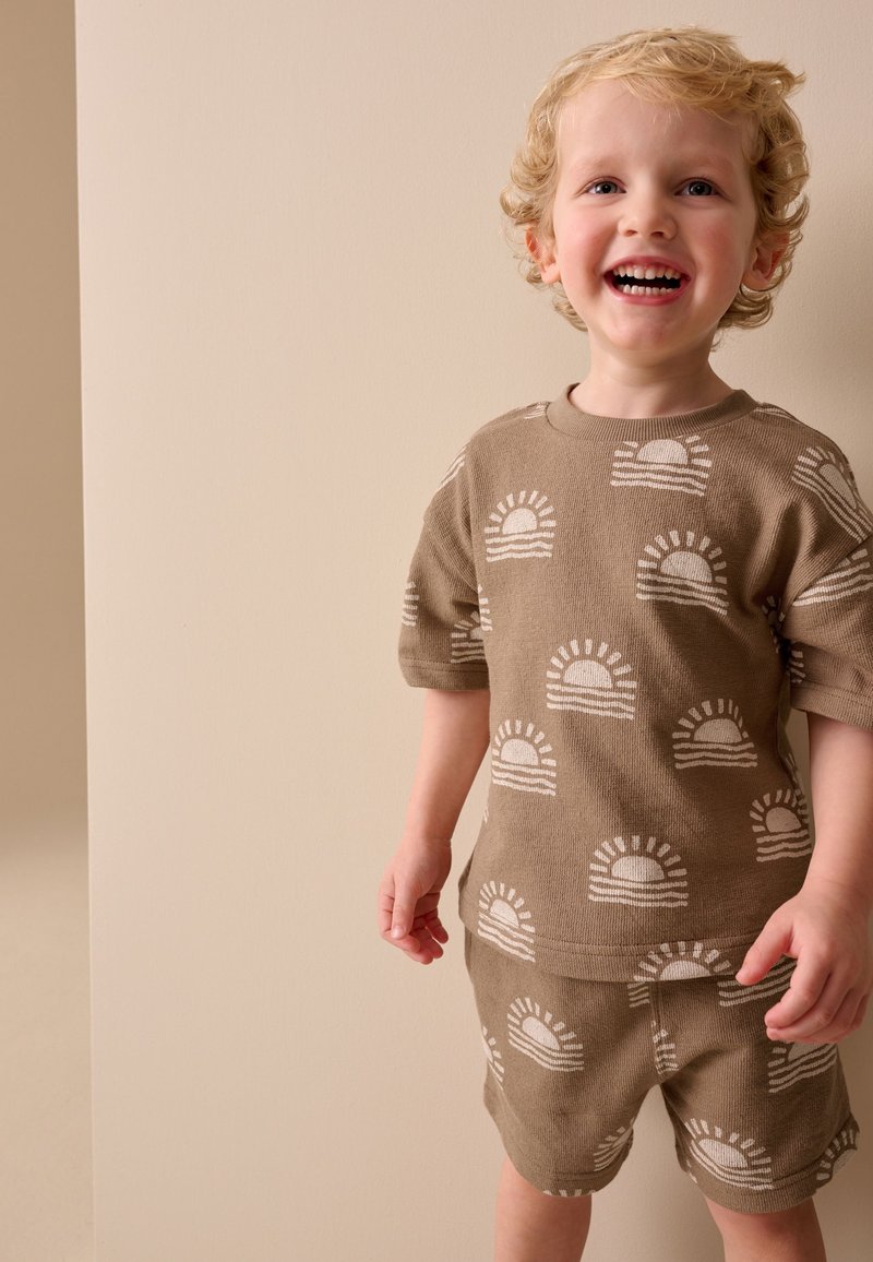 Niño pequeño sonriente con cabello rubio rizado lleva una camisa y pantalones cortos marrones a juego con patrones blancos de sol sobre un fondo beige.