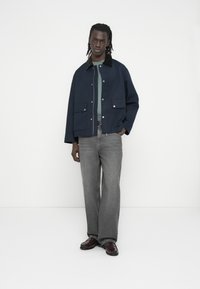 Veste bleu marine foncé avec col et poches avant, associée à un jean gris à jambes larges et des chaussures bordeaux. Design épuré et minimaliste.