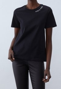 T-shirt en coton noir à manches courtes, col rond, avec broderie florale décorative près de l'épaule. Assorti à un pantalon noir ajusté.