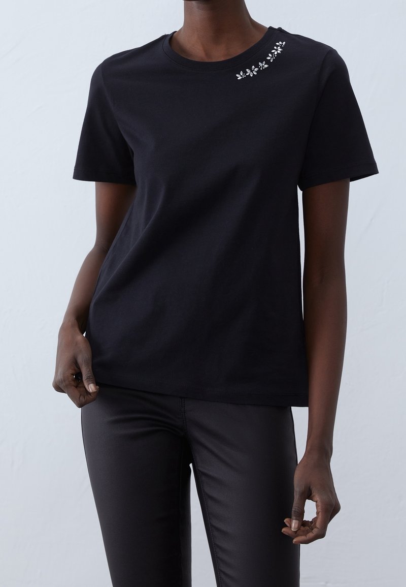 T-shirt en coton noir à manches courtes, col rond, avec broderie florale décorative près de l'épaule. Assorti à un pantalon noir ajusté.