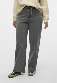 Jeans largi gri, din denim, cu talie cu șnur și textură subtilă. Asortați cu adidași negri și gri, pe un fundal alb.