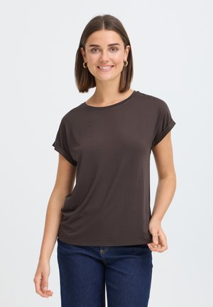 Jeune femme portant un haut marron foncé à manches courtes et un jean bleu, souriante avec des cheveux courts et raides et des boucles d'oreilles en or.