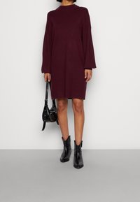 Burgunderfarbenes Strickkleid in knieumspielender Länge mit langen Ärmeln und hohem Kragen, kombiniert mit schwarzen Knöchelstiefeln und einer kleinen schwarzen Handtasche. Glatte Textur.