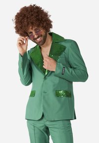 Costume vert avec un col en sequins brillants et des rabats de poches ; pantalon assorti ; mannequin portant des lunettes de soleil rondes et une perruque bouclée brune.