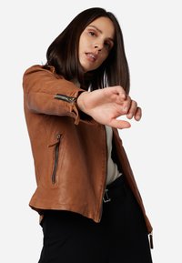 Braune Lederjacke mit tailliertem Design, ausgestattet mit Reißverschlusstaschen und langen Ärmeln. Die Textur ist glatt mit sichtbaren Nähten.