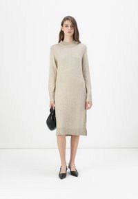 Beige kötött ruha kerek nyakkal, hosszú ujjakkal, oldalsó hasítékokkal és bordázott véggel; fekete táskával és hegyes orrú fekete cipőkkel párosítva.