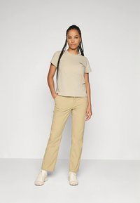 Beżowy t-shirt z krótkim rękawem i beżowe spodnie, o luźnym kroju, zestawione z jasnymi trampkami, na neutralnym tle.