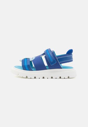 Modra sandal z dvema nastavljivima trakoma; ima belo debelo podplat in teksturirano površino. Vidne svetlo modre notranje obrobe.