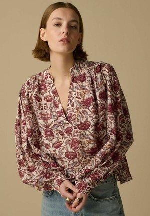 Femme aux cheveux courts bruns portant un chemisier floral à manches longues bouffantes et un jean bleu clair sur un fond beige.