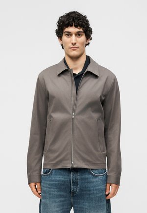 Muž s kučeravými vlasmi, ktorý má na sebe sivú bundu na zips cez čierne tričko a modré džínsy, stojaci pred jednofarebným svetlým pozadím.