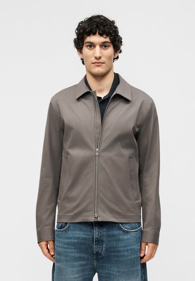 Homme aux cheveux bouclés portant une veste grise à fermeture éclair sur un t-shirt noir et un jean bleu, debout devant un fond clair uni.