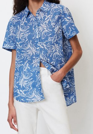 Personne portant une chemise bleue à manches courtes avec imprimé floral blanc et un pantalon blanc, une main dans la poche.