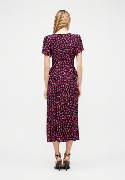 Γυναίκα με κυματιστά ξανθά μαλλιά που φοράει navy midi φόρεμα με κόκκινο φλοράλ μοτίβο και κοντά μανίκια, συνδυασμένο με μαύρα σανδάλια με ψηλό τακούνι, από πίσω.