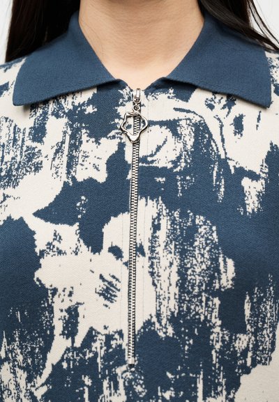 Κοντινό πλάνο πουκάμισου με μοτίβο σε ναυτικό μπλε και λευκό, με γιακά σε ναυτικό μπλε και ασημένιο φερμουάρ στο κέντρο, σε σχήμα τριφυλλιού με τέσσερα φύλλα.