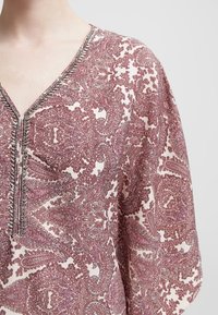 Blusa a fiori paisley in bordeaux e crema con scollo a V, caratterizzata da motivi intricati e filo argentato lungo il panneggio.