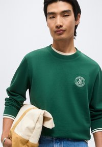 Νέος άνδρας που φοράει πράσινο φούτερ με λογότυπο σε λευκό, κρατά μια τσάντα σε μπεζ και καφέ χρώματα, στέκεται σε απλό λευκό φόντο.