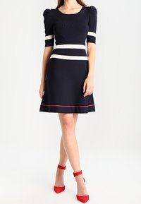 Femme portant une robe bleu marine à rayures blanches et rouges, manches courtes bouffantes, associée à des escarpins rouges à bride autour de la cheville, posant devant un fond blanc.