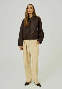 Koperkleurige leren cropped jas met een kraag en ritssluiting, gecombineerd met lichtbeige geplooide broeken en donkerbruine puntige schoenen.